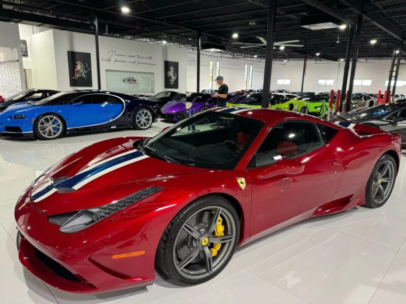 A red 458 Speciale with white and blue stripes gleams in the showroom, surrounded by other luxury cars, including a blue and a green model. This stunning lineup of modern Ferraris dazzles under the bright lights of the well-lit garage.