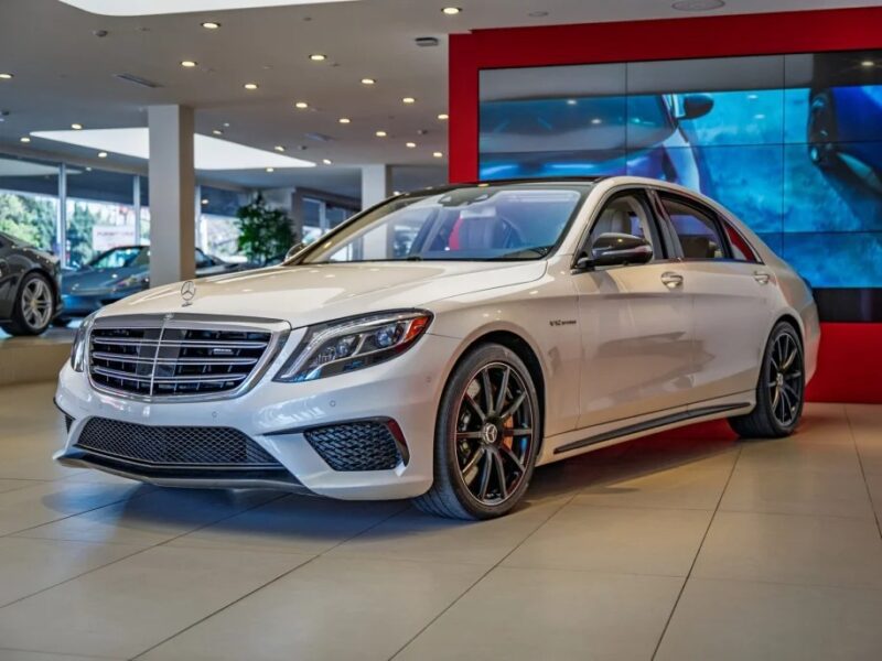 A white luxury sedan, exuding elegance, is displayed in a showroom with a red and white interior design. Large windows and modern lighting accentuate its sophisticated allure. Discover this blend of affordable luxury sedans now for sale.