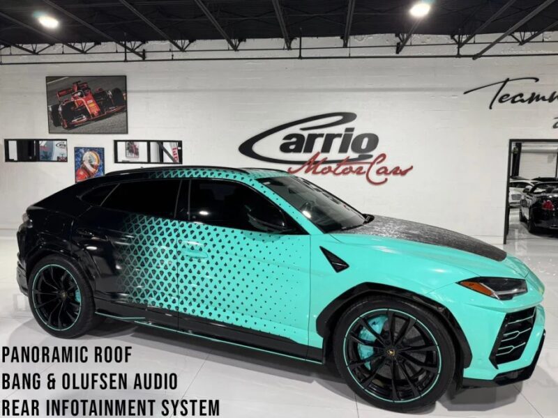 A Lamborghini Urus with a turquoise and black geometric pattern is parked indoors. This luxury SUV features a panoramic roof, Bang & Olufsen audio, and a rear infotainment system, offering an unparalleled driving experience. Now available for sale to discerning buyers.