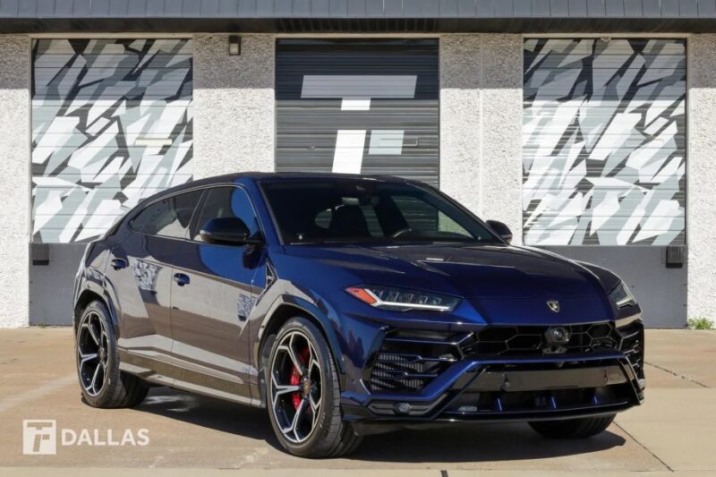 Urus Lamborghini Biru yang menakjubkan, salah satu SUV mewah untuk dijual, diparkir di depan gedung dengan pintu garasi berpola dan kata "Dallas" di sudut kiri bawah.