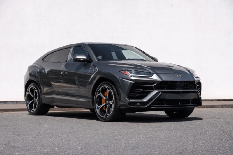 The sleek dark gray Lamborghini Urus SUV is elegantly parked on the asphalt against a crisp white wall, showcasing its exquisite design. If you're searching SUVs for sale with unmatched style and performance, this model stands out as one of the best.