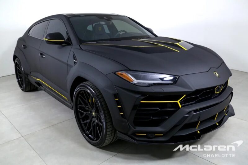 Matte black Lamborghini Urus with yellow accents and a sleek design features a prominent front grille. Displayed in the showroom, the "McLaren Charlotte" logo graces the corner, highlighting one of the finest Lamborghini SUVs for sale today.