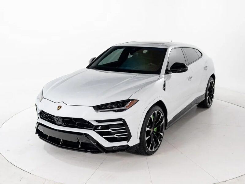 The Lamborghini Urus, a white luxury SUV with sleek design, black accents, and large alloy wheels, is elegantly showcased indoors against a white background. Experience one of the best SUVs for sale that seamlessly blends performance and style.