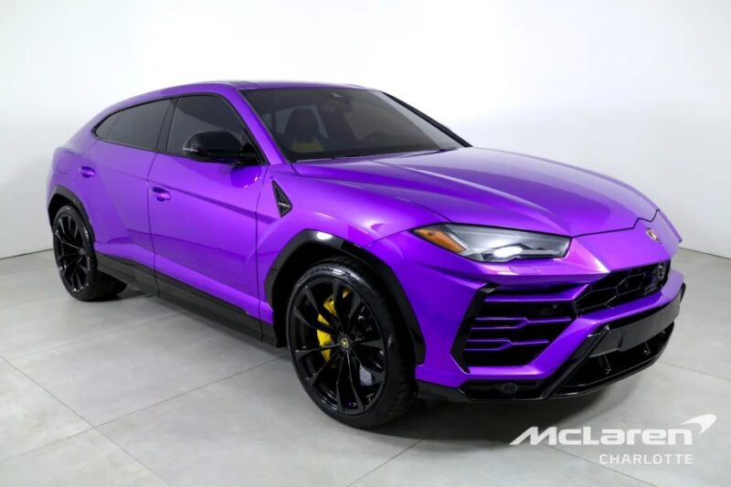 A purple Lamborghini Urus SUV with sleek black wheels is prominently displayed in a well-lit showroom, captivating potential buyers with its unique allure, and is now available for sale.