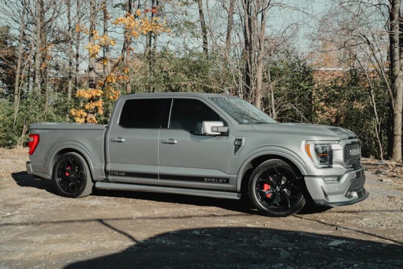 A gray Shelby Ford F-150 pickup truck is parked outdoors on a dirt surface, with trees and sparse foliage in the background—a standout among Shelby Trucks for sale.