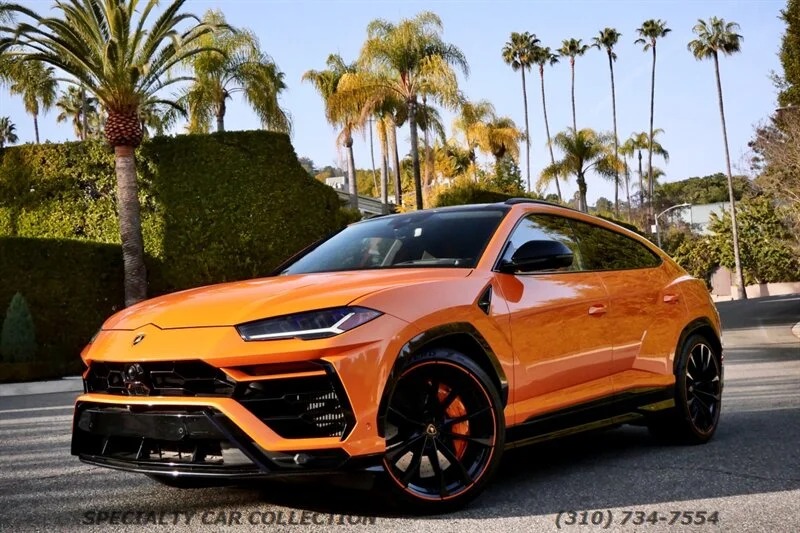 The best Lamborghini, a vibrant orange Lamborghini Urus, is parked majestically in front of swaying palm trees on a sunlit street.