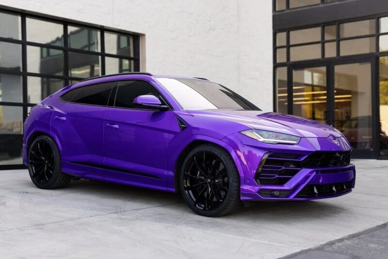 A striking purple Lamborghini Urus SUV is parked in front of a building with large glass windows, presenting a perfect blend of style and power.