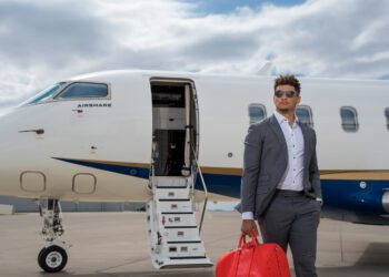 A person in a gray suit and sunglasses stands holding a red bag near a private jet with an open door on a cloudy day, epitomizing the pinnacle of private aviation.