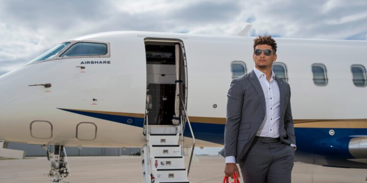 A person in a gray suit and sunglasses stands holding a red bag near a private jet with an open door on a cloudy day, epitomizing the pinnacle of private aviation.