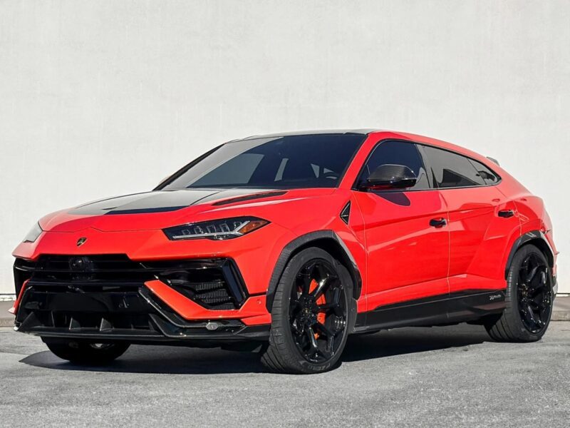 A striking red and black Lamborghini Urus sports SUV stands proudly on a concrete surface, viewed from the front and side against a plain white wall. Perfect for those seeking the best Lamborghini, this Urus is among the top SUVs for sale today.