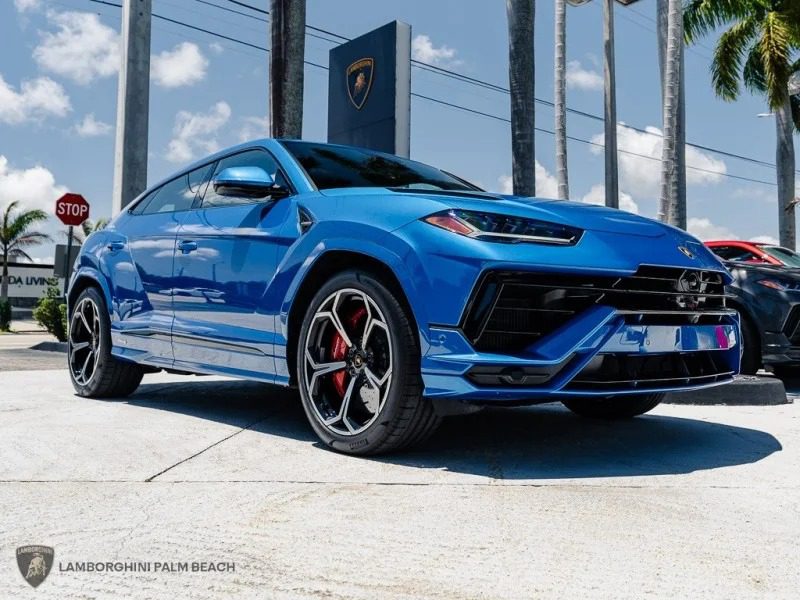A blue Lamborghini Urus, one of the best Lamborghini SUVs for sale, is parked outdoors near a dealership, with palm trees and a Lamborghini sign in the background.