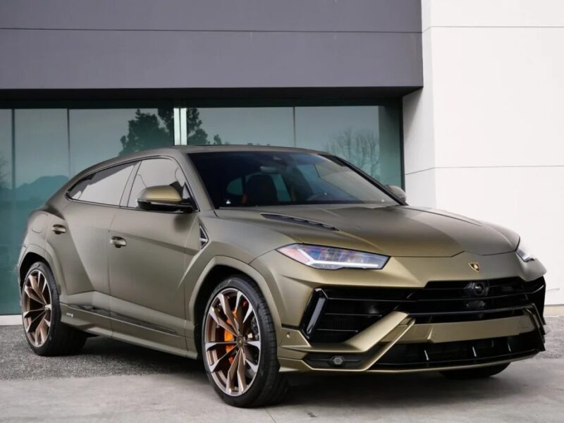 A matte green Lamborghini Urus with an angular design, large wheels, and tinted windows is parked outside a modern building. This striking SUV exudes luxury and performance, combining sleek lines with a commanding presence. It's the perfect vehicle for those seeking adventure and style.