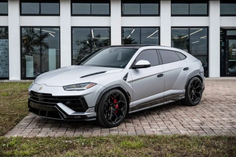 A silver luxury Lamborghini Urus is parked on a brick driveway in front of a building with large windows. This sleek SUV, featuring tinted windows and black wheels, is now for sale.