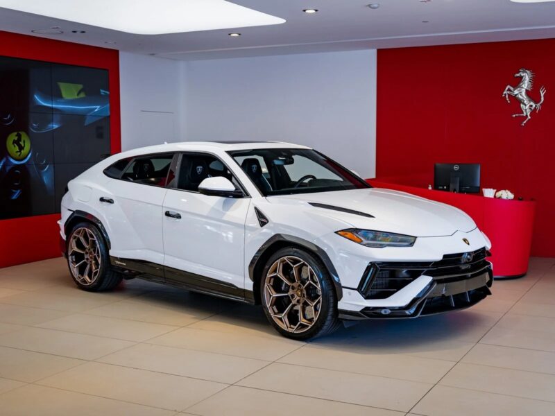 A white Lamborghini Urus, one of the best Lamborghini SUVs for sale, is parked inside a modern showroom with red walls and a Ferrari logo in the background.