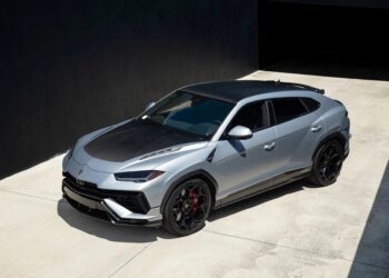 A silver Lamborghini Urus, one of the best Lamborghinis, with black accents and red brake calipers is parked on a concrete driveway beside a dark building.