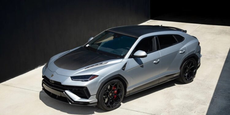A silver Lamborghini Urus, one of the best Lamborghinis, with black accents and red brake calipers is parked on a concrete driveway beside a dark building.
