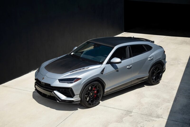 A silver Lamborghini Urus, one of the best Lamborghinis, with black accents and red brake calipers is parked on a concrete driveway beside a dark building.