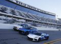 Three cars, including a sleek Chevrolet model, are parked on a racetrack in front of empty grandstands at the "World Center of Racing", capturing the essence of speed and innovation reminiscent of a NASCAR Prototype.