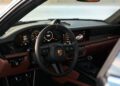 The luxurious interior of a Porsche 911 S/T features a black steering wheel proudly bearing the iconic Porsche logo, digital dashboard displays, and elegant brown leather accents—a true testament to the craftsmanship celebrated by RM Sotheby's.