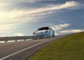 A sleek Porsche 911 S/T cruises along an asphalt road with a metal guardrail, set against a backdrop of partly cloudy skies and rolling grassy hills, reminiscent of scenes you'd find at a prestigious car auction.