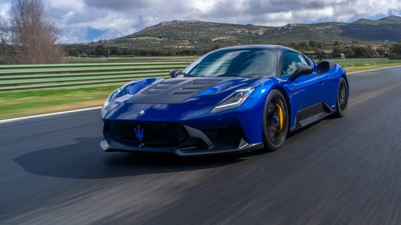 A blue sports car driving on a racetrack with mountains in the background under a cloudy sky.