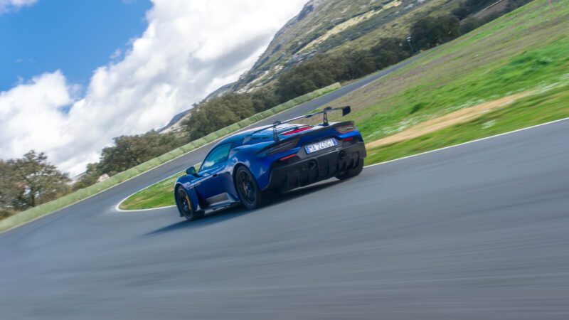 A blue sports car navigating a curve on a racetrack, with mountains and clouds in the background.