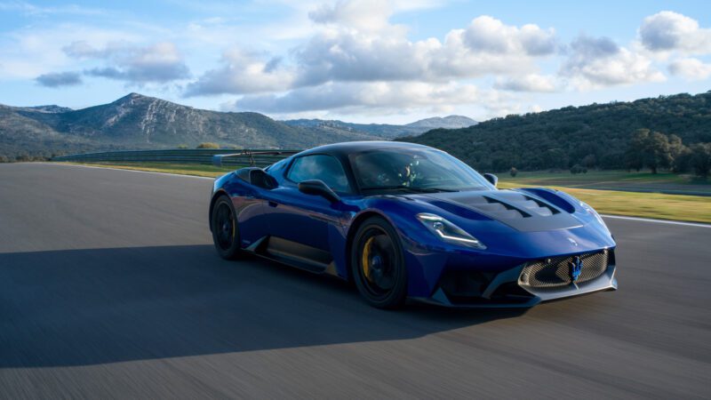 A blue sports car driving on a racetrack against a backdrop of mountains and cloudy sky.