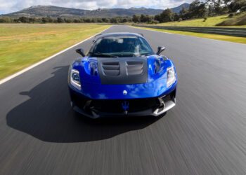 A blue sports car drives on a race track with mountains in the background under a partly cloudy sky.