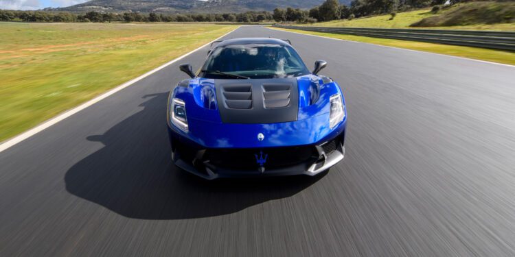A blue sports car drives on a race track with mountains in the background under a partly cloudy sky.