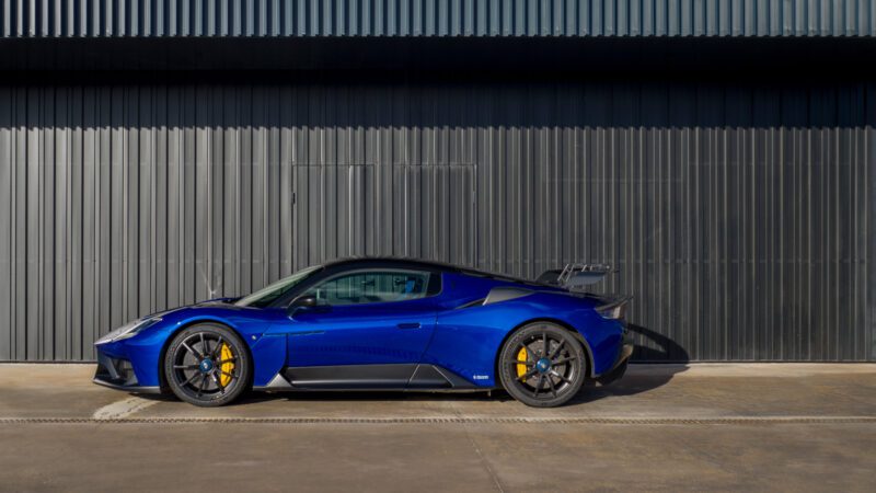 Blue sports car with yellow brake calipers parked beside a corrugated metal wall in the sunlight.