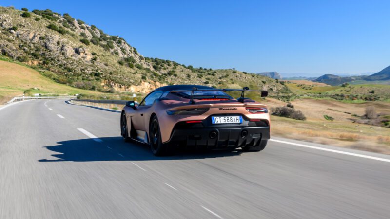 A sports car with a "Maserati" badge drives on a scenic road, surrounded by hills and clear blue sky.