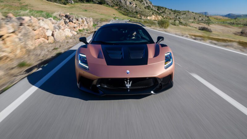 A copper-colored sports car speeds down a rural road, surrounded by greenery and hills.