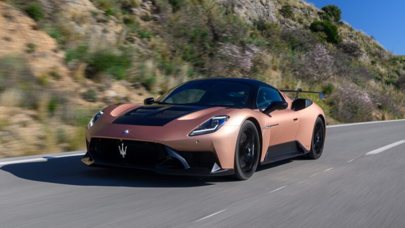 A copper-colored sports car drives on a sunny, mountainous road. The car has a sleek design with a prominent front grille and aerodynamic features.