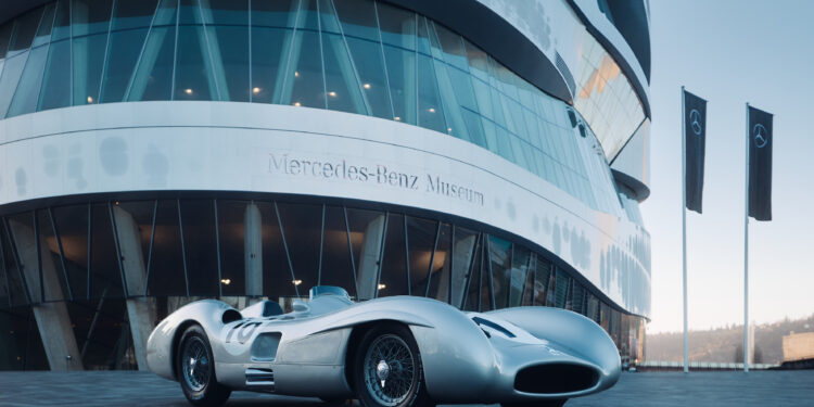A vintage silver Mercedes-Benz W 196 R race car is elegantly parked in front of the iconic Mercedes-Benz Museum, its architectural glass and metal facade providing a striking backdrop. A similar model once fetched $53.9M at an RM Sotheby's Auction, highlighting its prestigious legacy.
