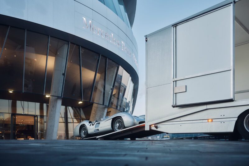 Sebuah mobil balap Mercedes-Benz klasik dengan hati-hati dimuat ke dalam trailer di luar bangunan kaca modern yang ramping dengan desain melengkung, memulihkan kenangan hari-hari kejayaannya di depan pelelangan RM Sotheby.