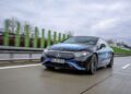 A blue Mercedes-Benz electric vehicle, powered by cutting-edge EV battery technology, glides on a wet highway against a backdrop of cloudy skies and green barriers.