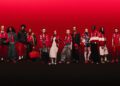 A group of 14 people in coordinated red-themed outfits stands in a row on a gradient red background, celebrating the 20th Anniversary of the Replica Collection. One person holds a checkered flag, embodying the spirit of PUMA x Scuderia Ferrari.