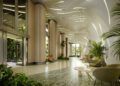 Welcome to the modern lobby of Tower 2, where a curved ceiling, tall columns, and expansive windows invite natural light. A green marble floor, sleek green chairs, lush plants, and wooden accents blend seamlessly to create a natural ambiance in the heart of Miami real estate.