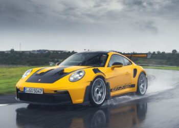 A yellow Porsche 992 911 GT3 RS zips along a wet road, its Michelin tires gripping the slick surface, while trees and buildings blur in the background.
