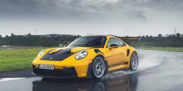 A yellow Porsche 992 911 GT3 RS zips along a wet road, its Michelin tires gripping the slick surface, while trees and buildings blur in the background.