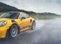 A yellow Porsche 992 911 GT3 RS with a rear wing speeds on a wet racetrack, spraying water. Hills and a cloudy sky add to the dramatic backdrop.