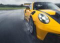 A yellow Porsche 992 911 GT3 RS with a black stripe speeds through a wet track, water splashing from its Wet-Weather Track Tires. The overcast sky looms in the background.