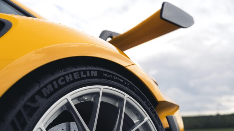 Close-up dari Porsche 992 911 GT3 RS kuning, fokus pada roda belakang dan spoiler. Ban berbunyi "Michelin Pilot Sport 3," dengan tetesan air berkilau di permukaannya yang ramping.