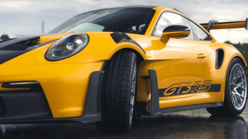 Porsche 911 GT3 RS kuning yang mencolok dengan aksen hitam ramping dan logo GT3 RS yang menonjol duduk di permukaan basah yang berkilau, menampilkan kecakapan ban lintasan cuaca basah yang mengesankan.