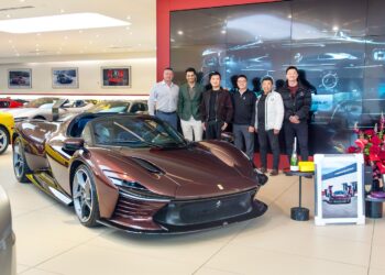 A group of six people stands behind a sleek, dark brown convertible in an upscale showroom filled with other luxury vehicles, including the impressive Ferrari Daytona SP3.