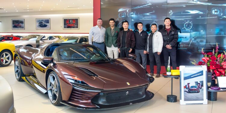 A group of six people stands behind a sleek, dark brown convertible in an upscale showroom filled with other luxury vehicles, including the impressive Ferrari Daytona SP3.