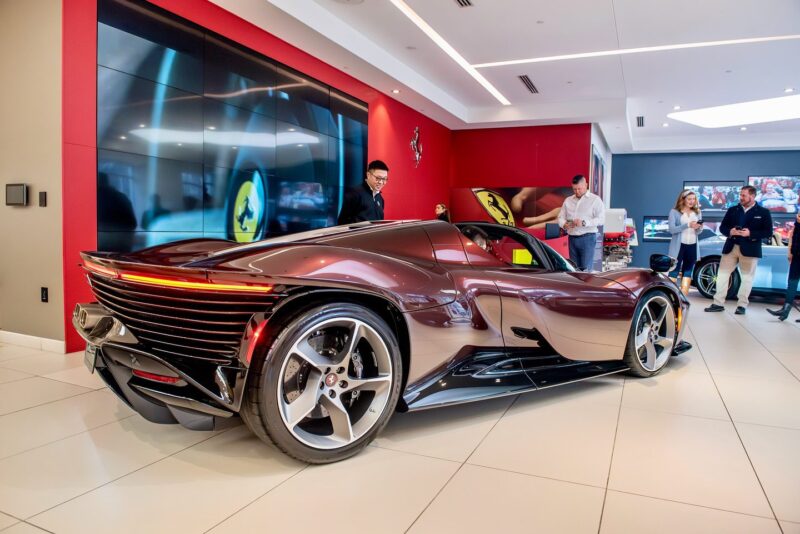 Hypercar coklat yang ramping, mungkin Daytona SP3 yang menakjubkan dari Ferrari, dipajang di ruang pamer. Pengamat yang antusias mengagumi mobil dengan latar belakang dinding merah dan putih dan layar besar.