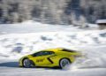 A yellow Lamborghini speeds through the snowy landscape of Livigno, kicking up snow in its wake. Snow-covered trees and a wooden cabin can be seen in the background, making it a thrilling snow driving experience.