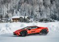 A red Lamborghini with "Accademia Neve" on the side zips down a snow-covered road, offering an exhilarating Snow Driving Experience. In the background, snow-laden trees and a cozy wooden cabin complete this picturesque scene in Livigno.