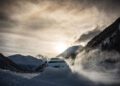 A Lamborghini car powers through the snow on a winding mountain road in Livigno, with the sun setting behind snowy peaks and a cloudy sky. This epic snow driving experience captures both thrill and serenity against a stunning alpine backdrop.